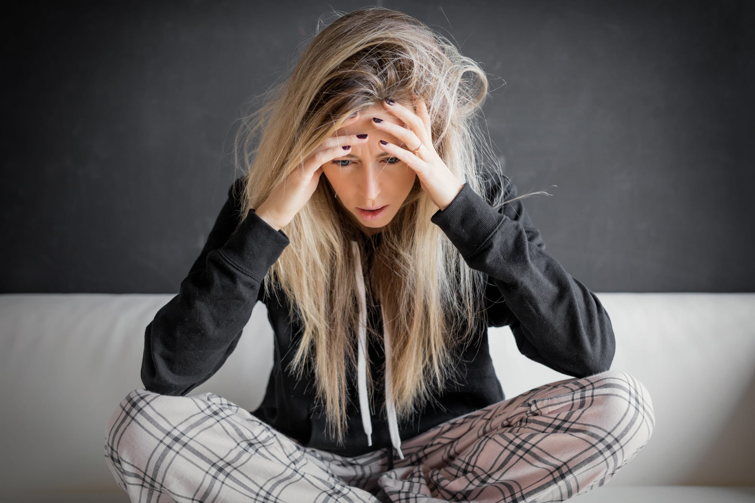 Woman sitting cross-legged on a couch, wearing plaid pants and a black hoodie, holding her head with both hands and looking down, appearing stressed or deep in thought.