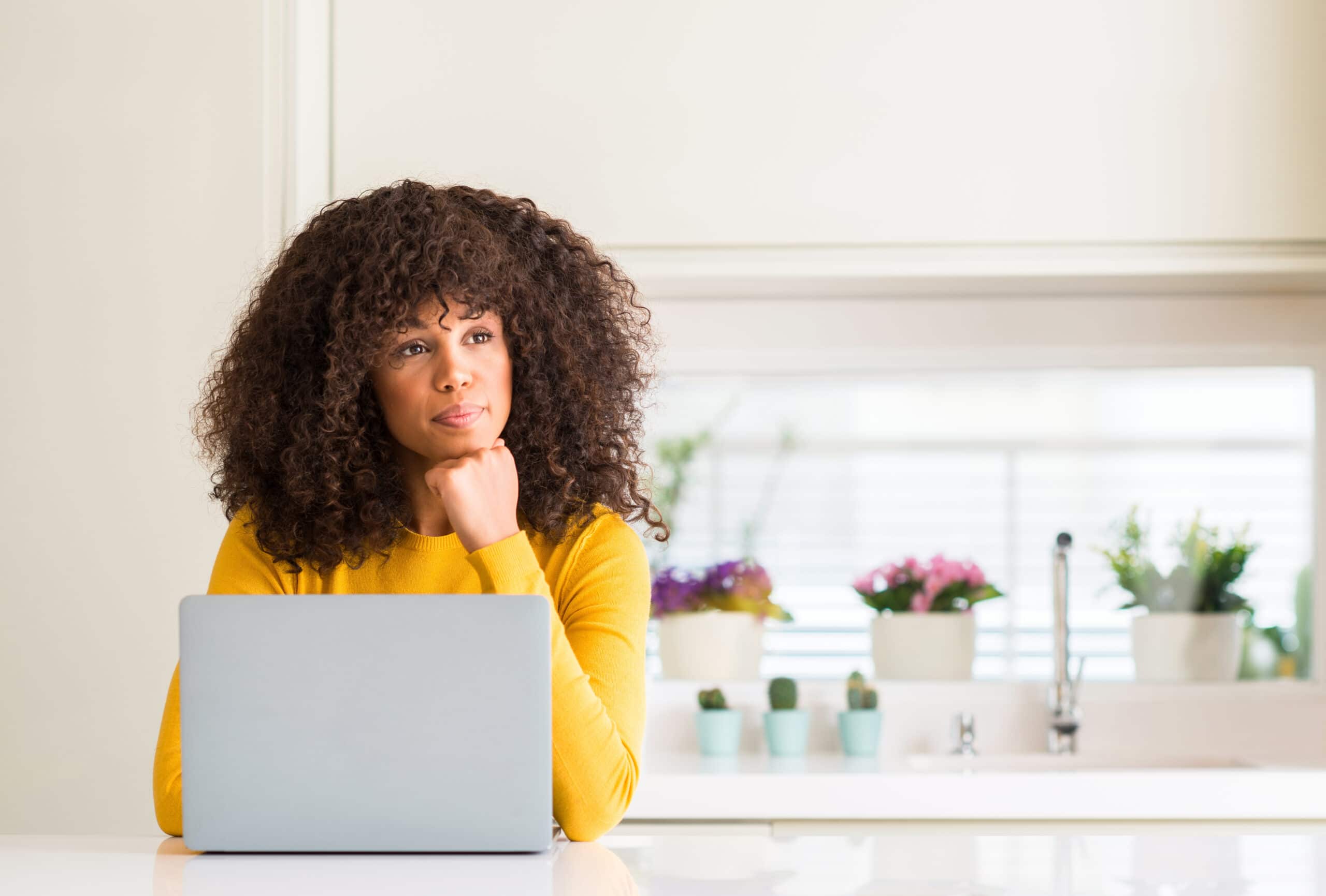 Woman with curly hair in a yellow top sits at a table with a laptop, resting her chin on her hand, in a bright kitchen with potted plants in the background.