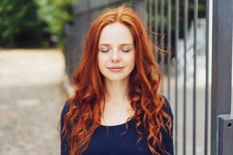 A woman with long red hair and fair skin stands outdoors with her eyes closed, wearing a navy blue top. A metal fence and blurry greenery are in the background.