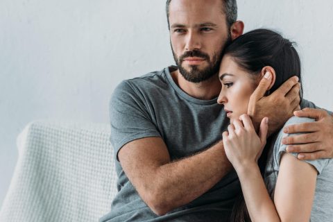 A man sits on a couch holding and comforting a woman who looks distressed, both wearing grey shirts.