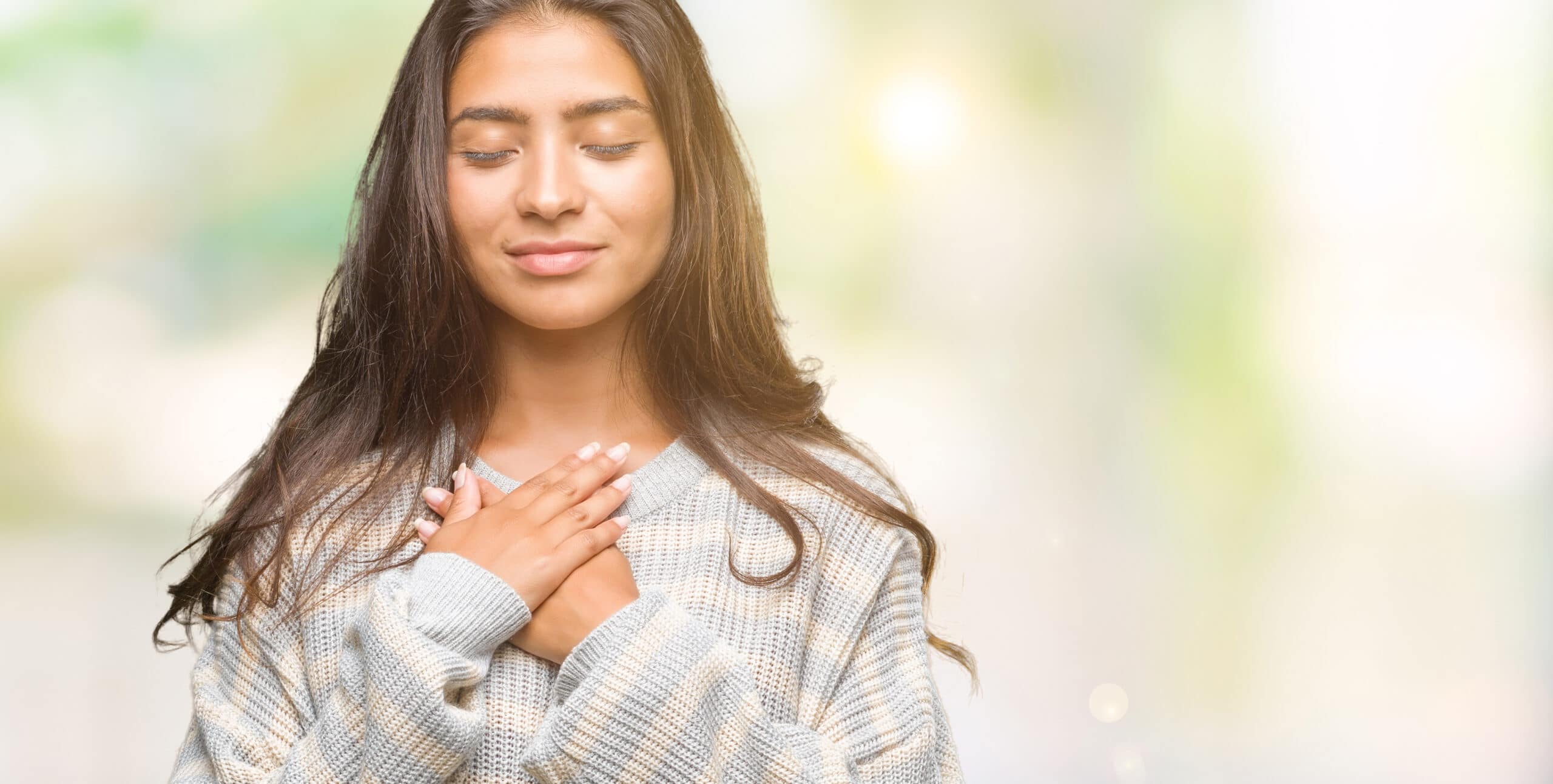 A woman with long brown hair stands with her eyes closed and both hands placed over her chest, wearing a light gray sweater against a blurred background.