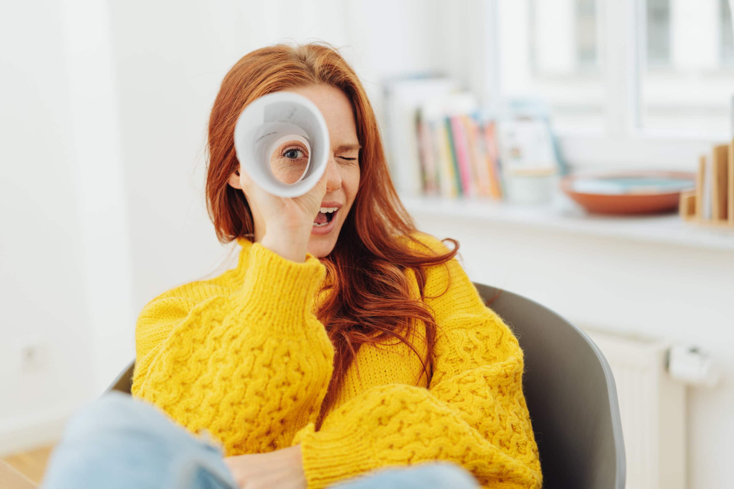 A person with long red hair in a yellow sweater looks through a rolled-up paper as if it were a telescope, sitting indoors with shelves and books in the background.
