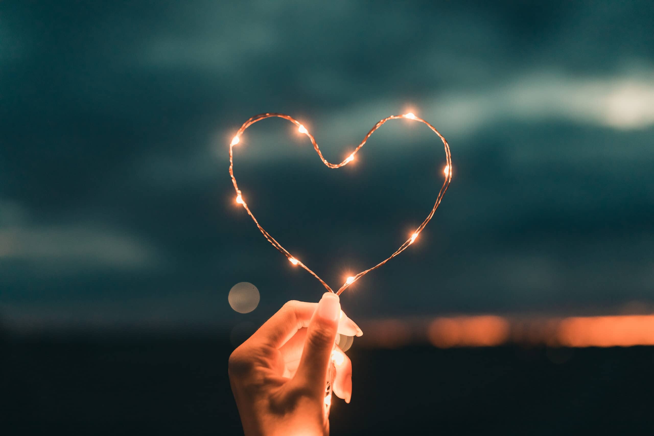 A hand holds a string of small lights shaped into a heart, with a blurred dark sky and distant lights in the background.