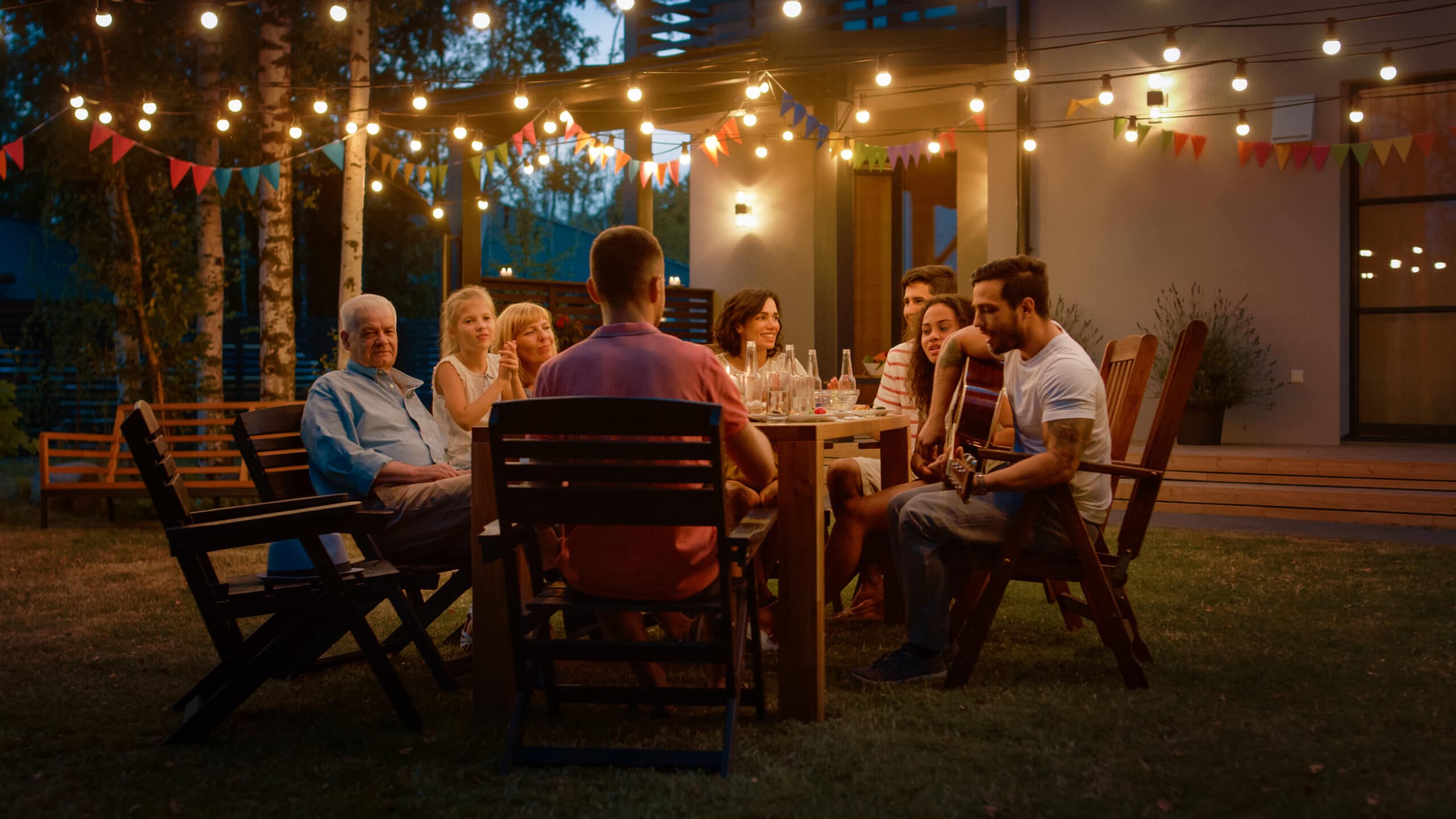 A group of people sit around a table outdoors in the evening, under string lights, with one person playing guitar.
