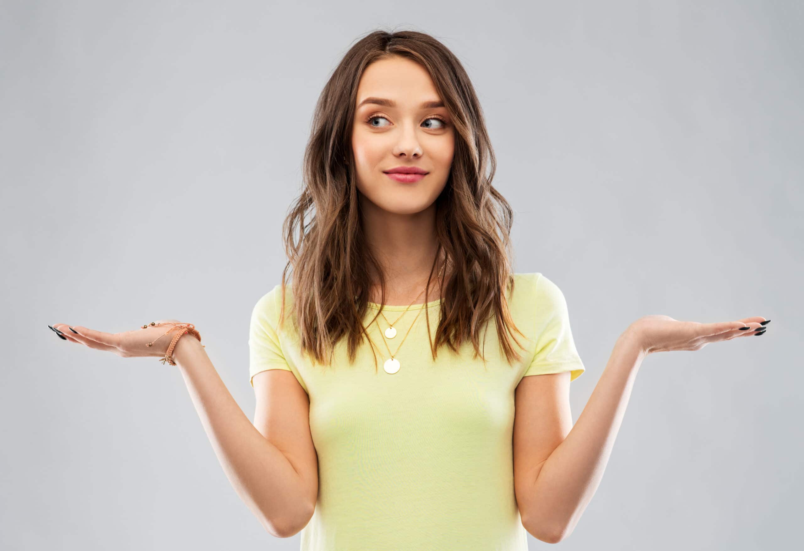 Young woman in a yellow shirt stands against a plain background, holding both hands up with palms facing upwards in a questioning or comparing gesture.