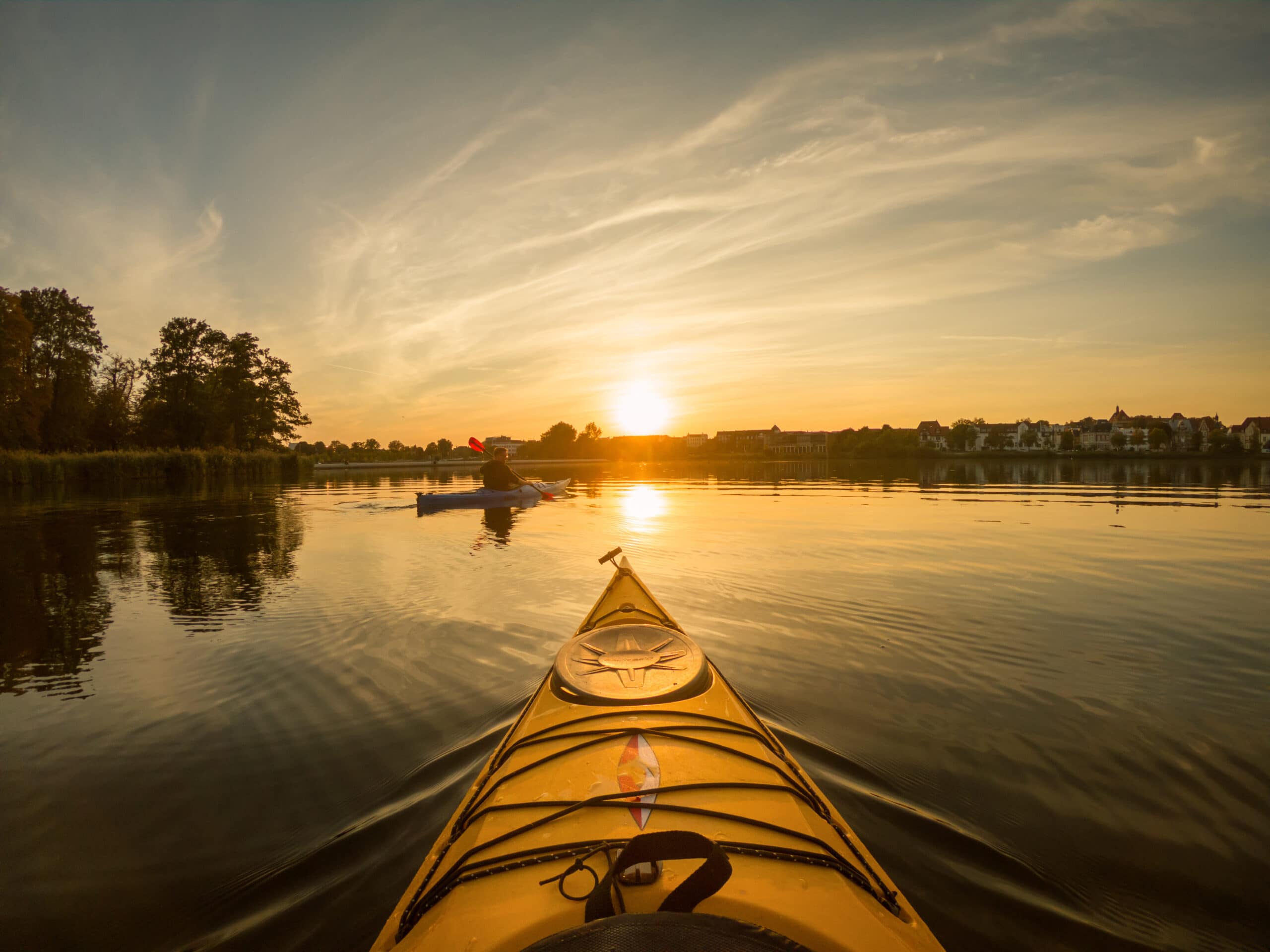 View from a yellow kayak on calm water at sunset, with another person kayaking in the distance, trees and buildings along the shoreline.