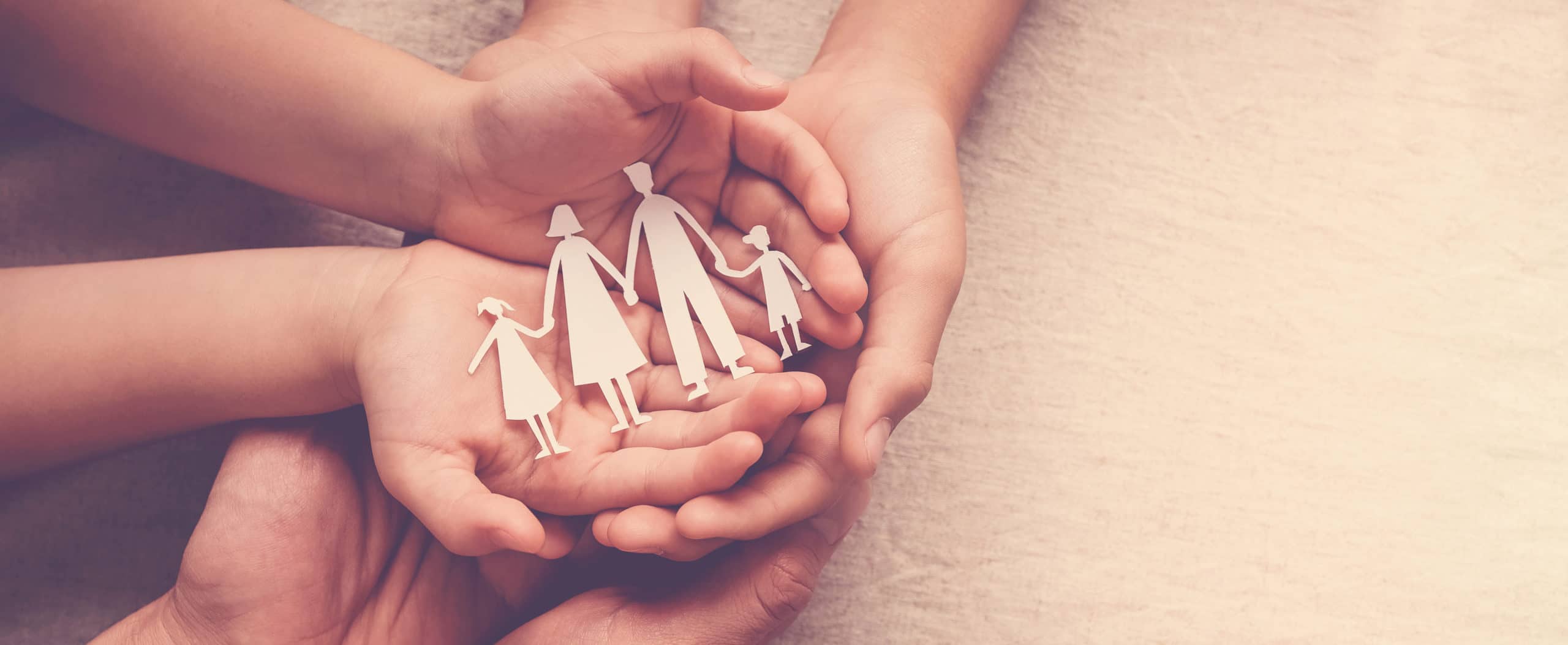 Two hands hold paper cutouts of a family of four, with two smaller hands resting on top, symbolizing family support and protection.