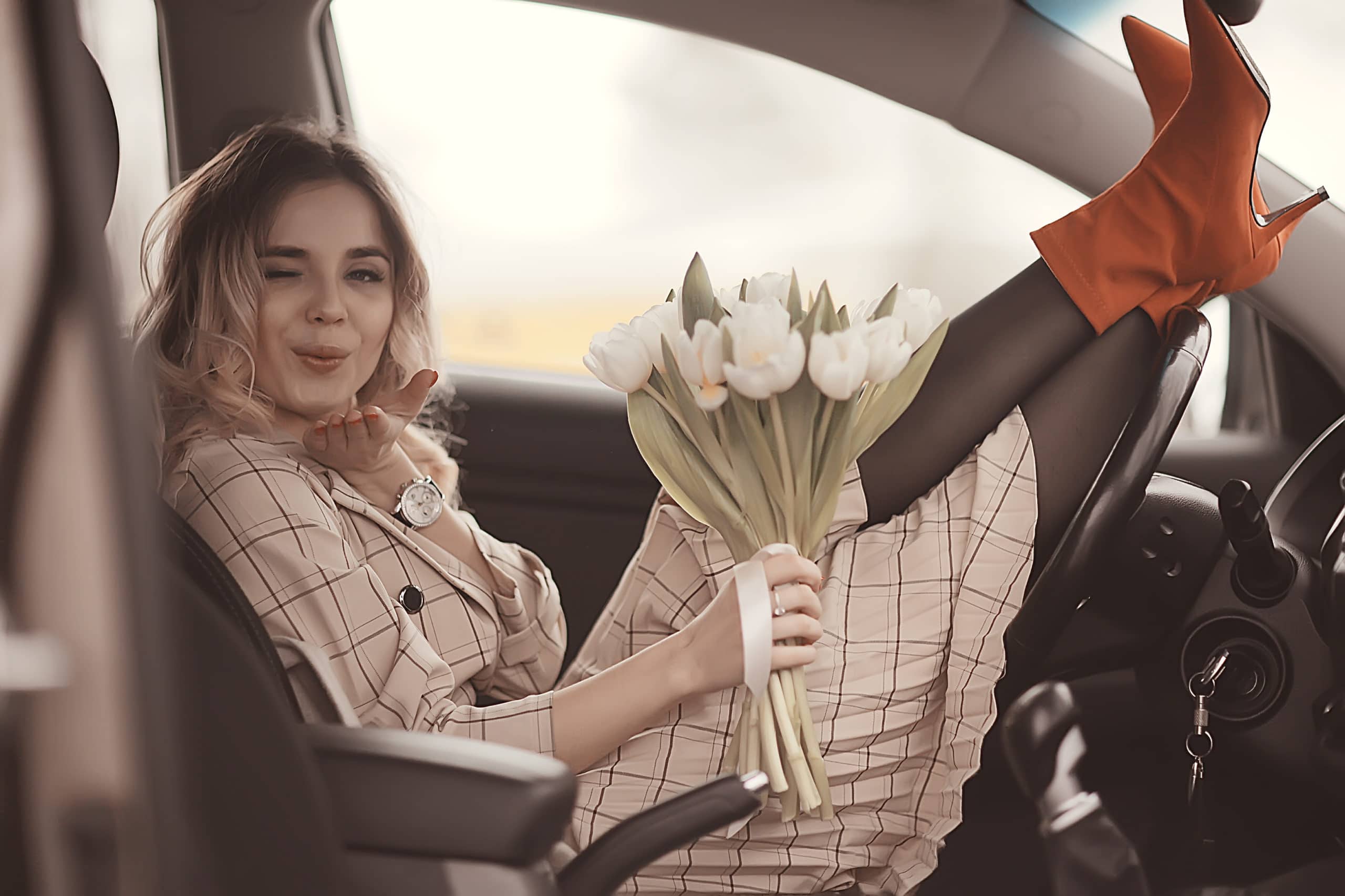 Woman sitting in a car with one leg on the dashboard, holding a bouquet of white tulips, wearing a plaid coat and red boots, blowing a kiss and winking.