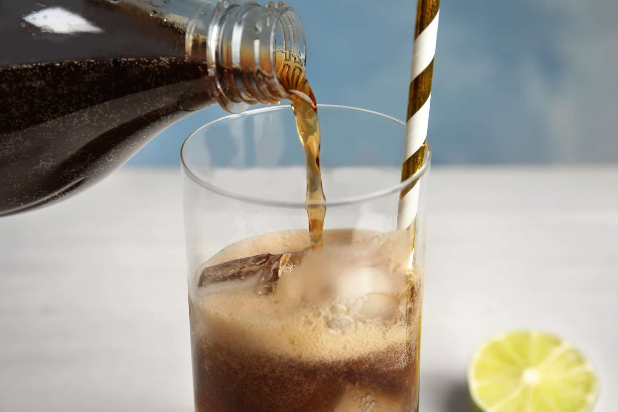 A glass of soda with ice is being filled from a bottle; a striped straw is in the glass and a lime wedge is on the table nearby.