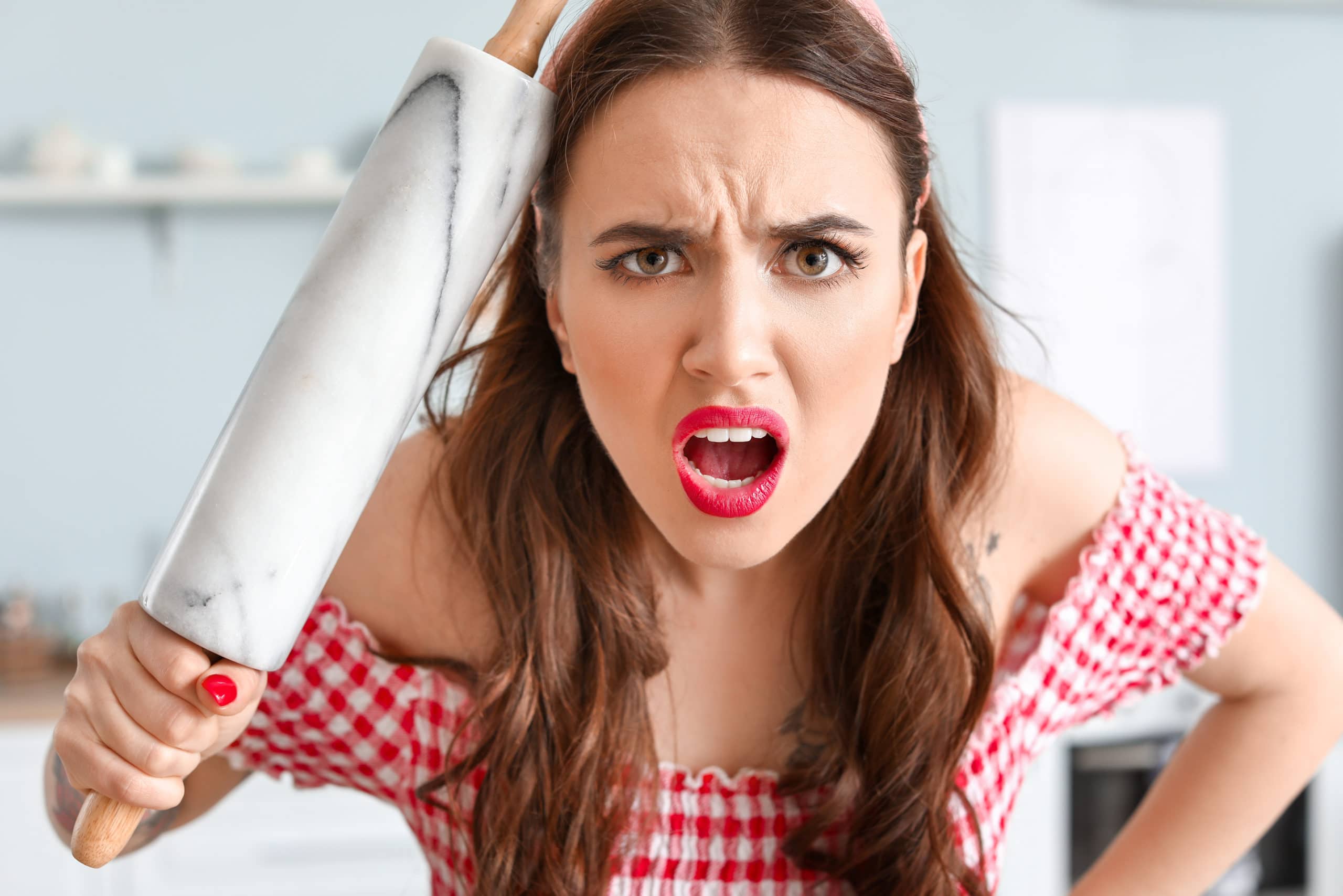 A woman in a red checkered top holds a marble rolling pin and looks directly at the camera with an angry, surprised expression.