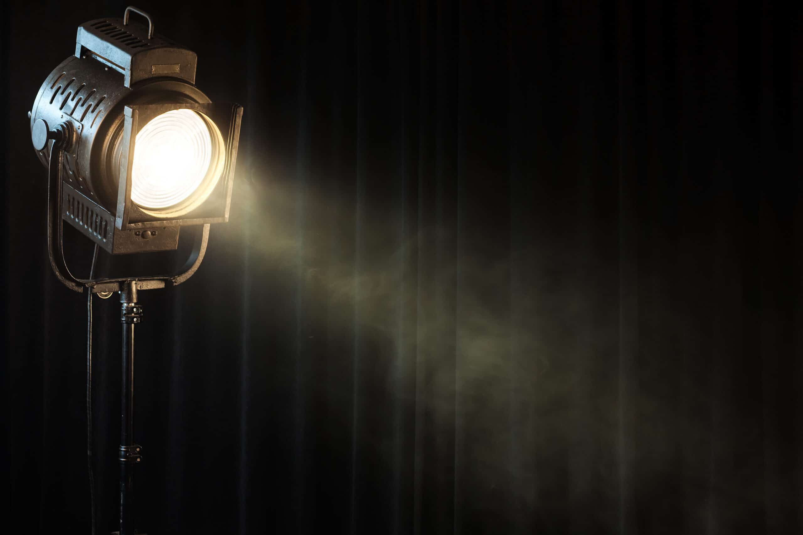 A stage spotlight is turned on, emitting a bright beam of light through mist in front of dark curtains.