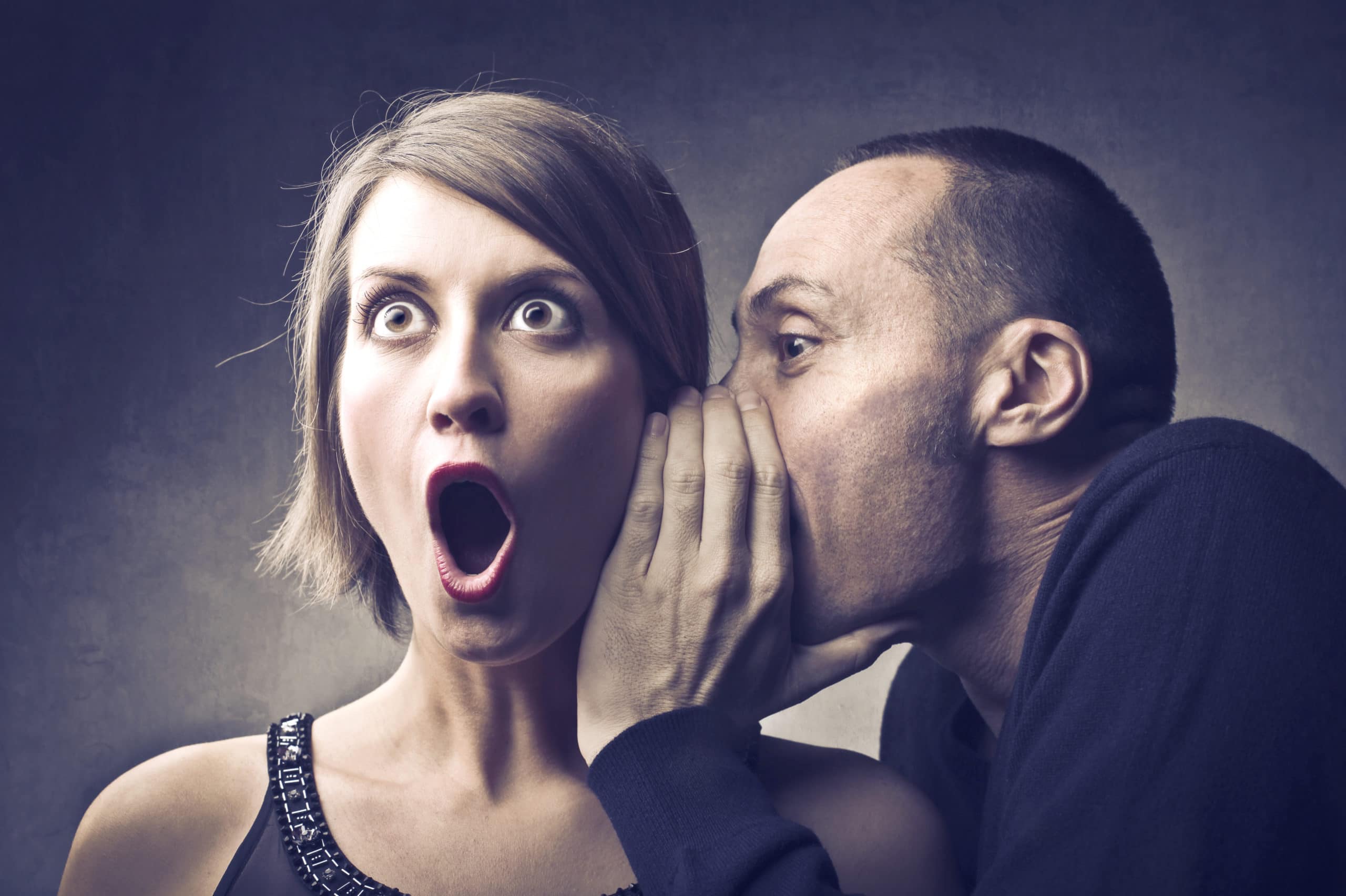 A man whispers into a woman’s ear; she looks surprised with wide eyes and an open mouth.