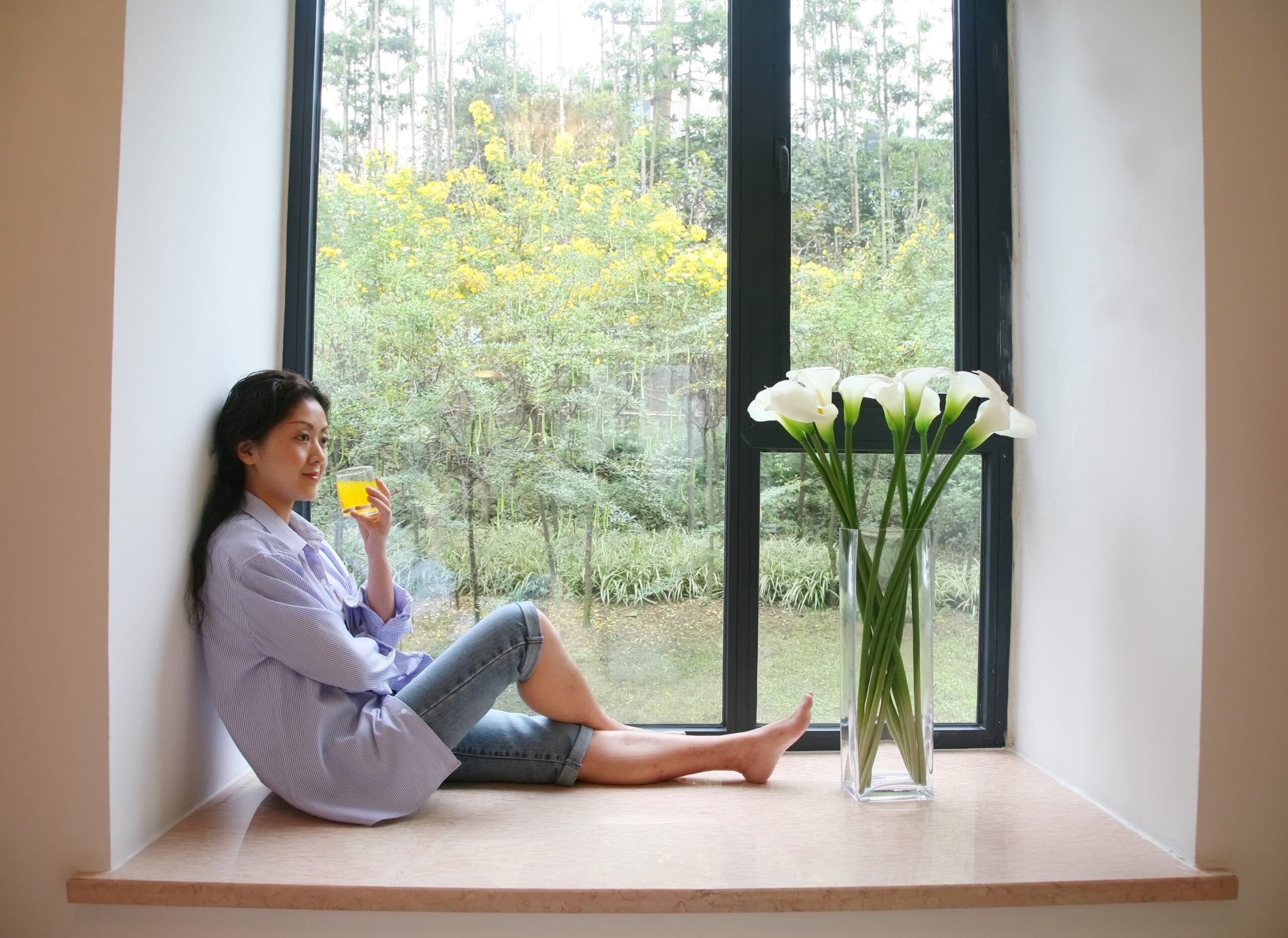 A woman sits on a windowsill holding a drink, with a large vase of white flowers beside her and a view of trees and yellow flowers outside the window.