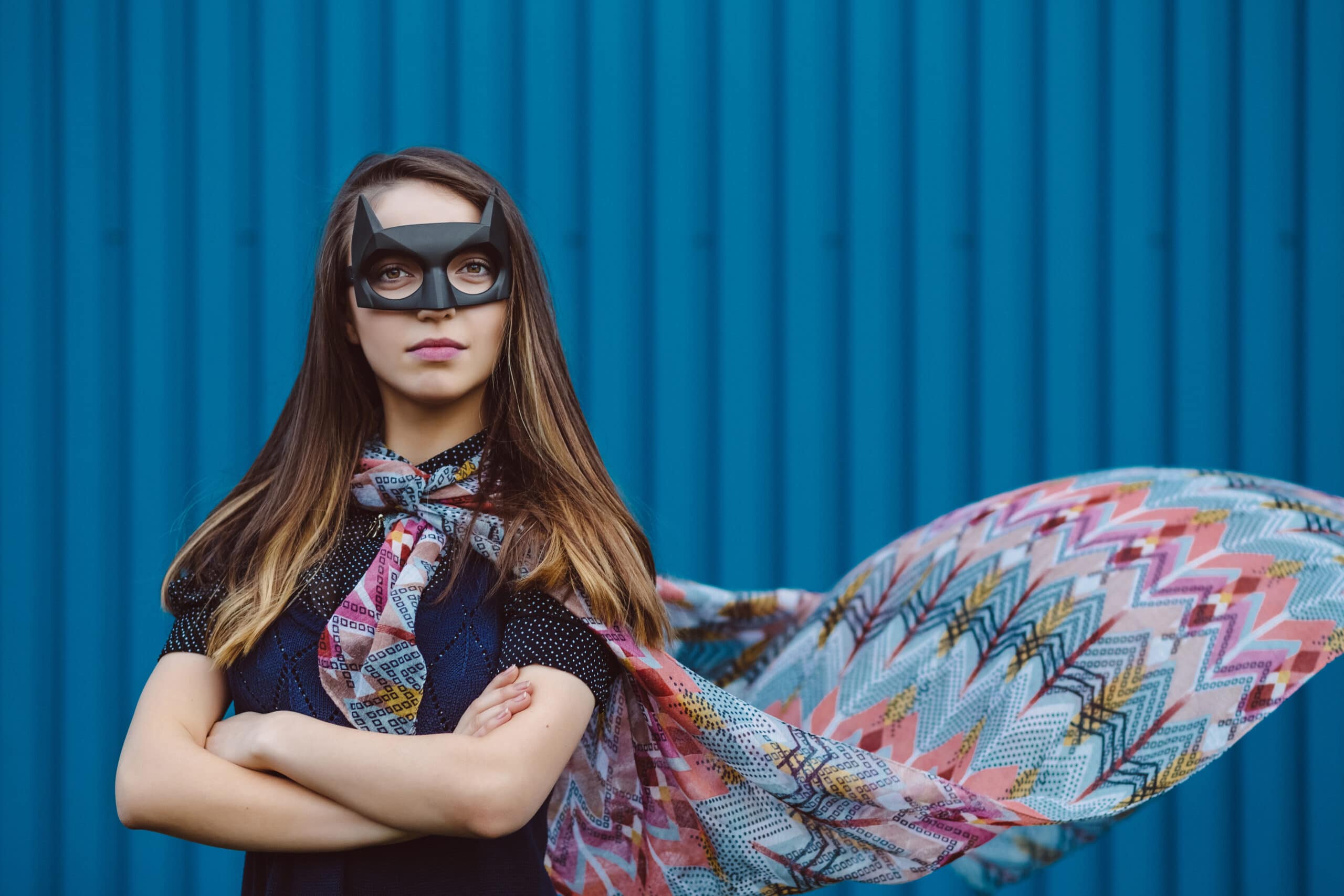 A woman wearing a black eye mask and multicolored cape stands with arms crossed in front of a blue corrugated metal wall.