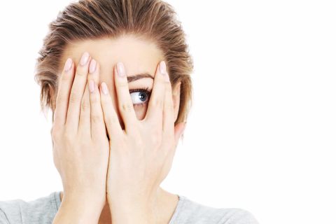 A person with light brown hair covers most of their face with their hands, peeking through their fingers with one visible blue eye against a white background.