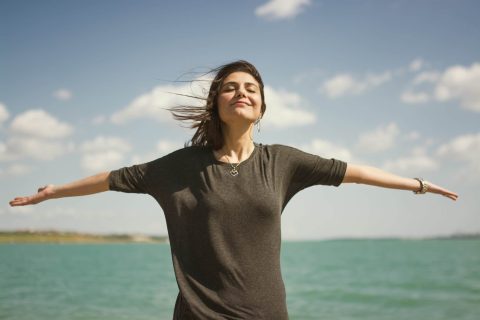 Woman standing near a body of water with arms outstretched, eyes closed in relief as the wind blows her hair under a partly cloudy sky.