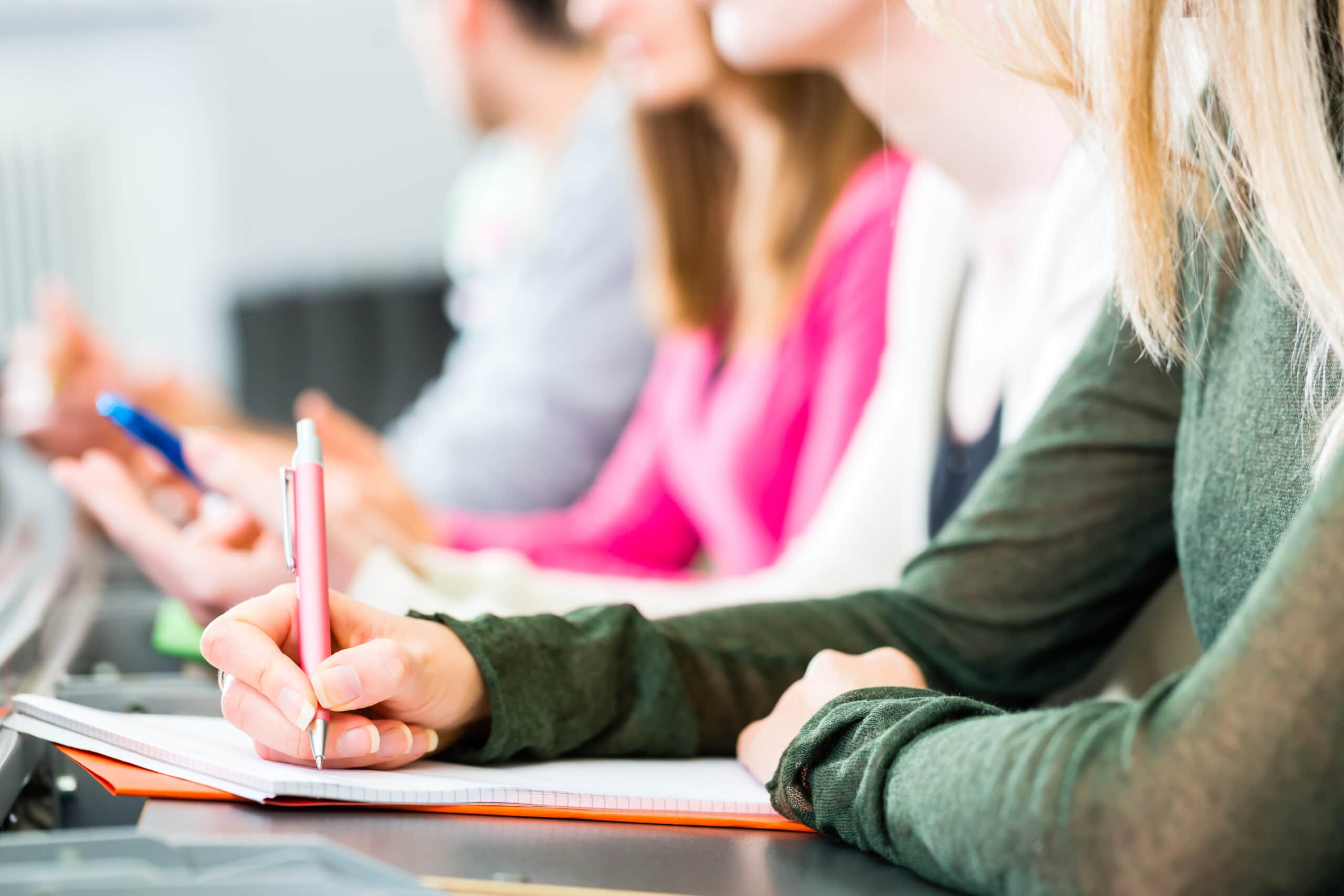 Several people sit in a row, focused on taking notes in notebooks with pens during what appears to be a class or lecture.