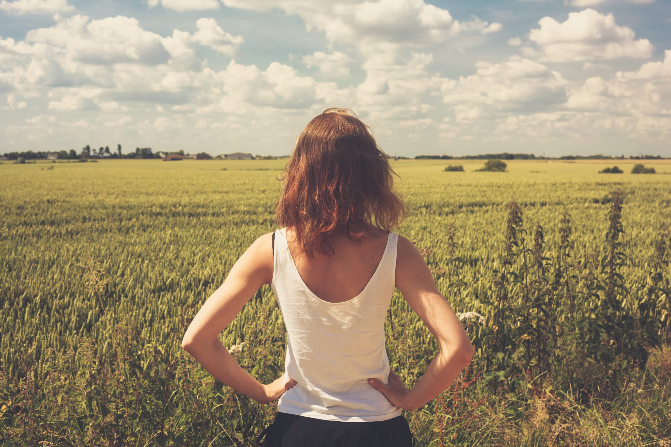 A person with shoulder-length hair stands with hands on hips, facing a vast green field under a partly cloudy sky.