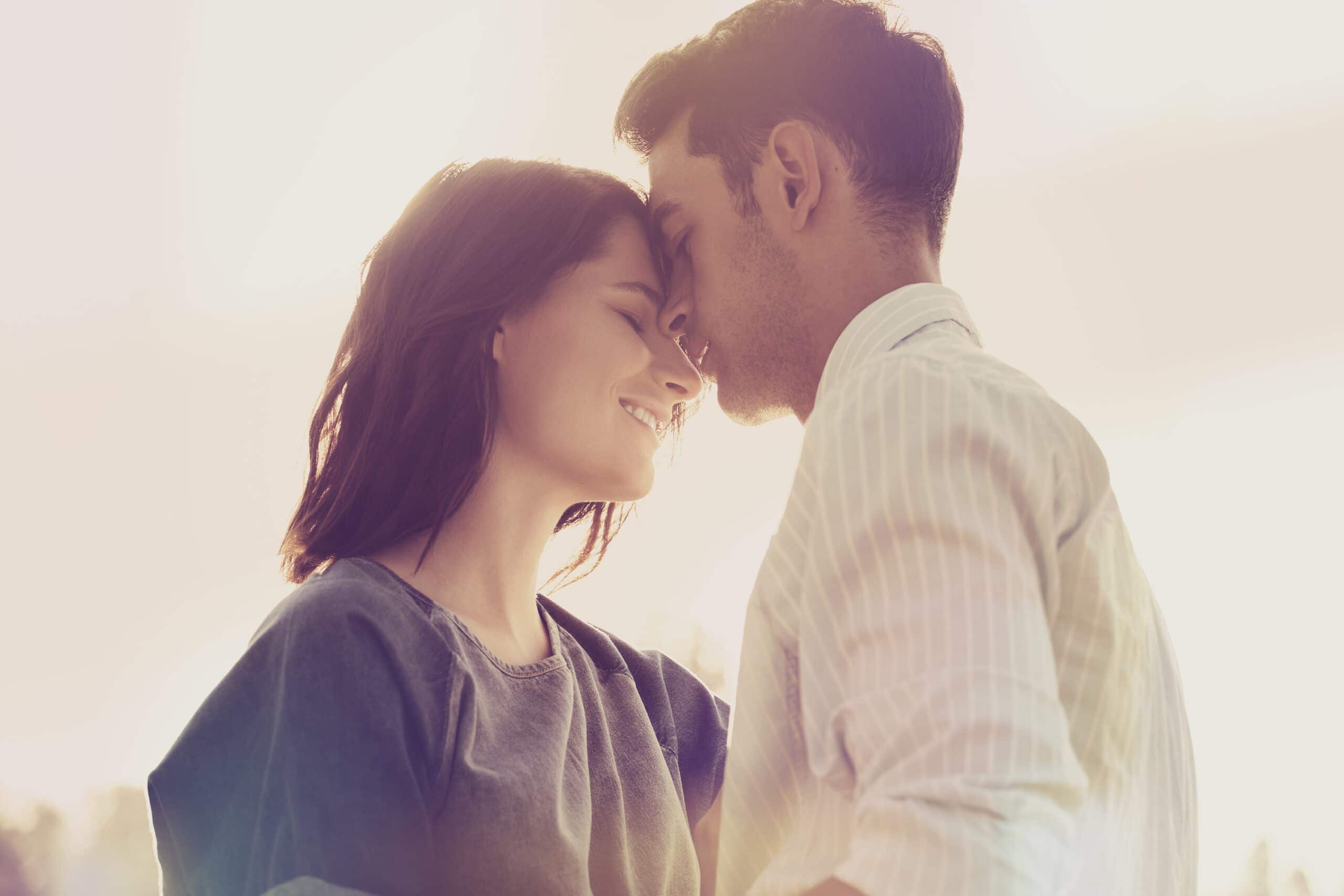 A man gently kisses a woman's forehead while she smiles, both standing close together in soft, natural light.