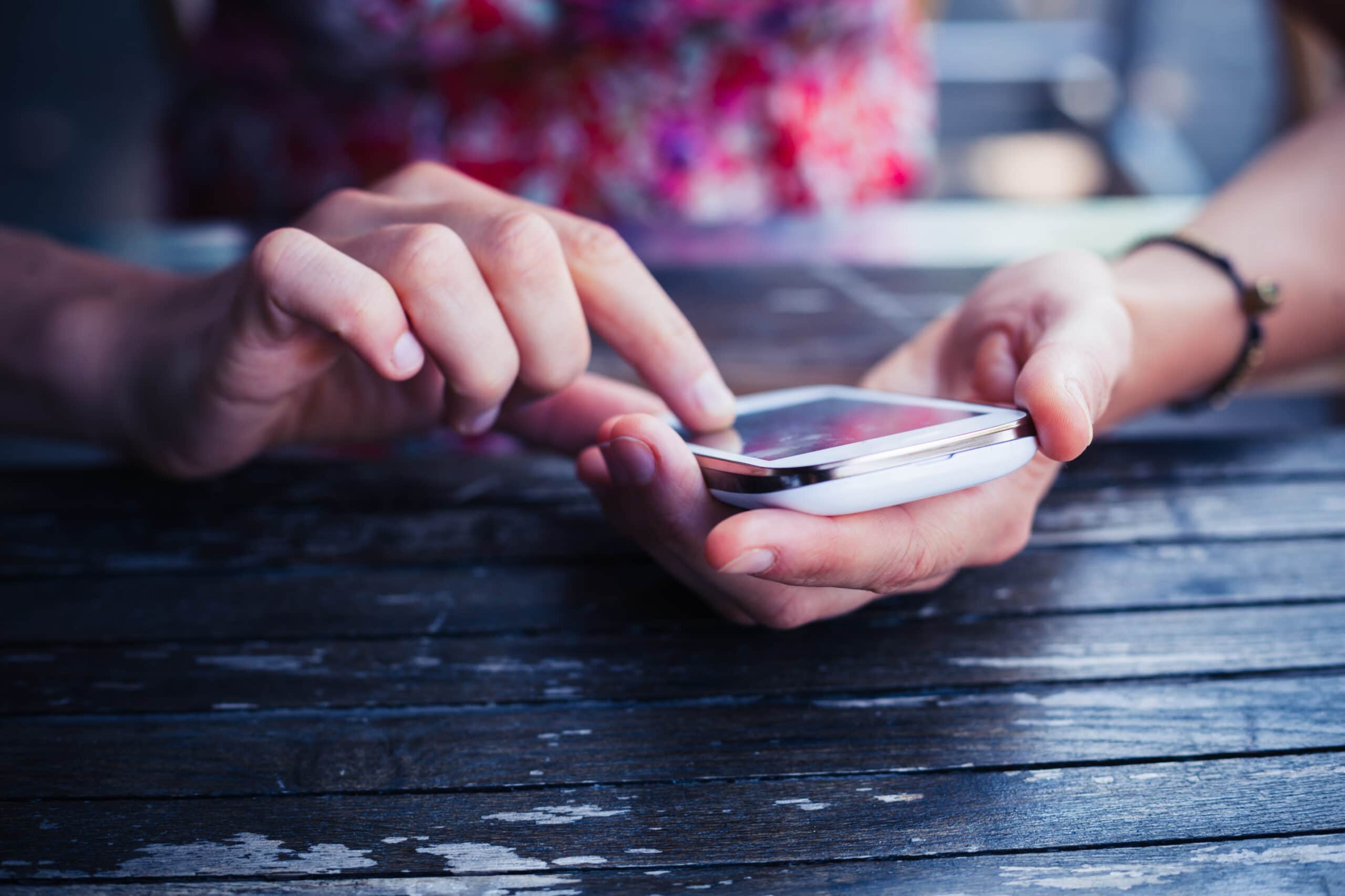 A person uses their right index finger to tap on a smartphone held in their left hand, sitting at a wooden table.