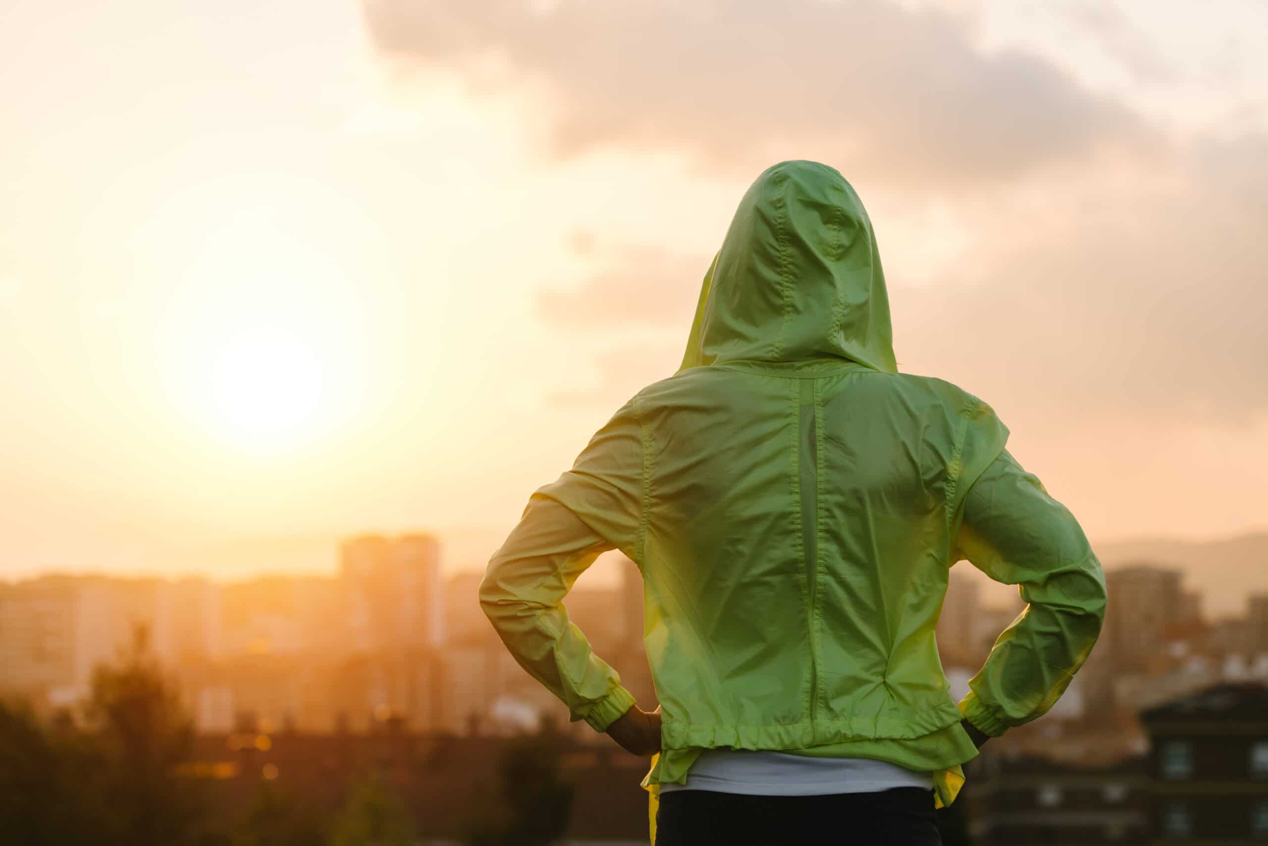Person in a green hooded jacket stands with hands on hips, looking at a cityscape during sunrise or sunset.