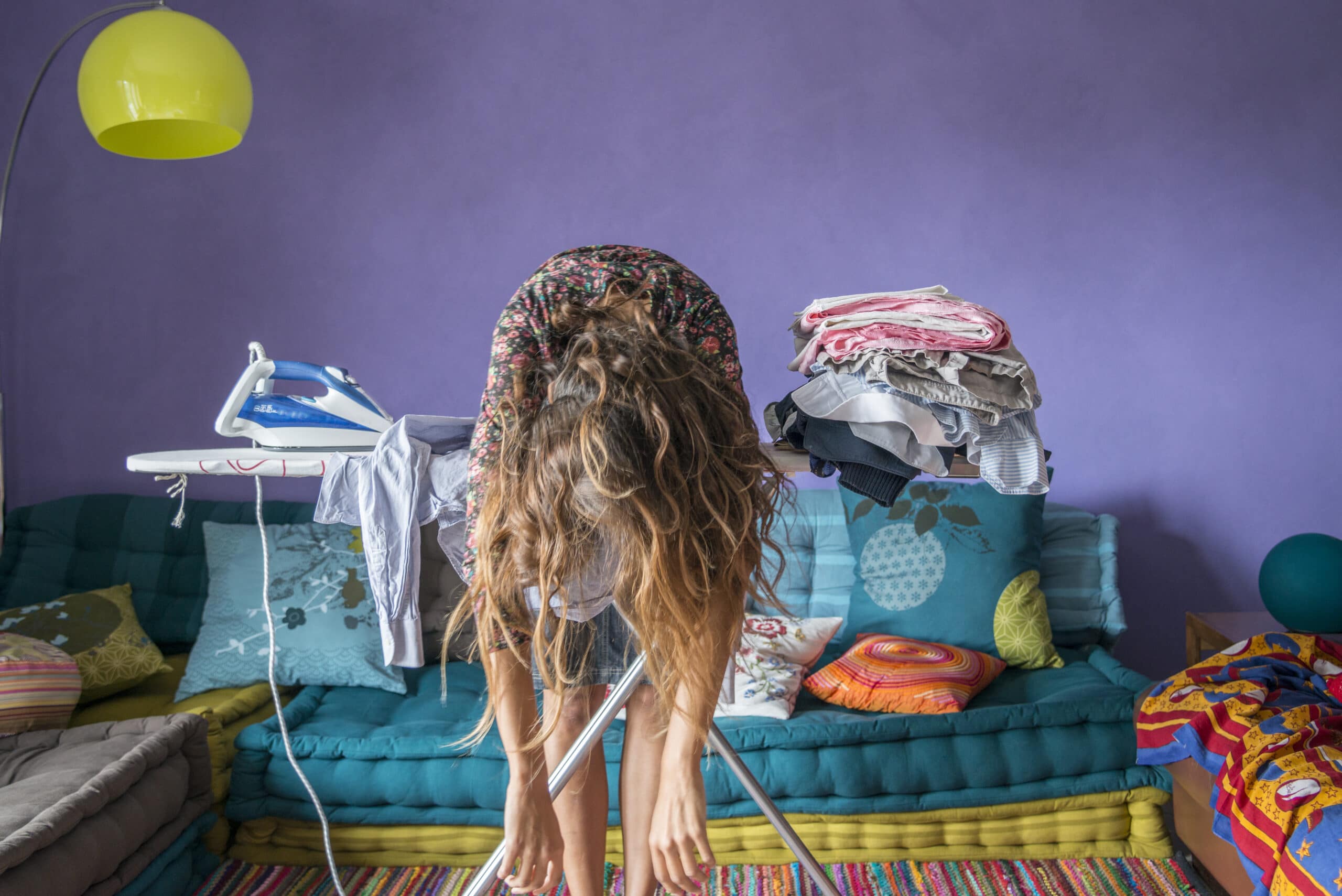 A woman leans over an ironing board, hair hanging down, surrounded by folded laundry in a colorful, cluttered living room.