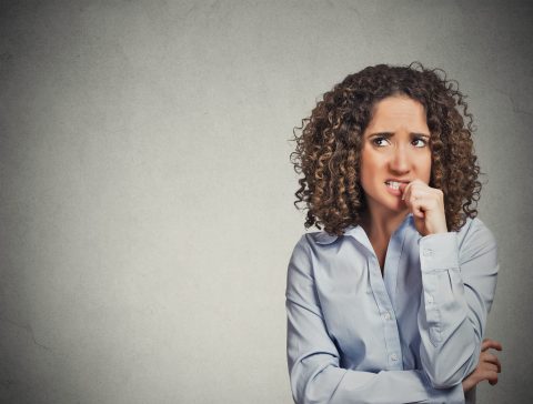 A woman with curly hair and a light blue shirt stands against a plain background, looking anxious and biting her fingernail.