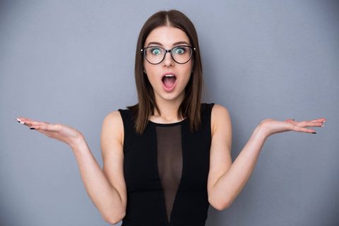 Woman with straight brown hair and glasses wearing a black sleeveless top, holding her hands up and looking surprised against a plain gray background.