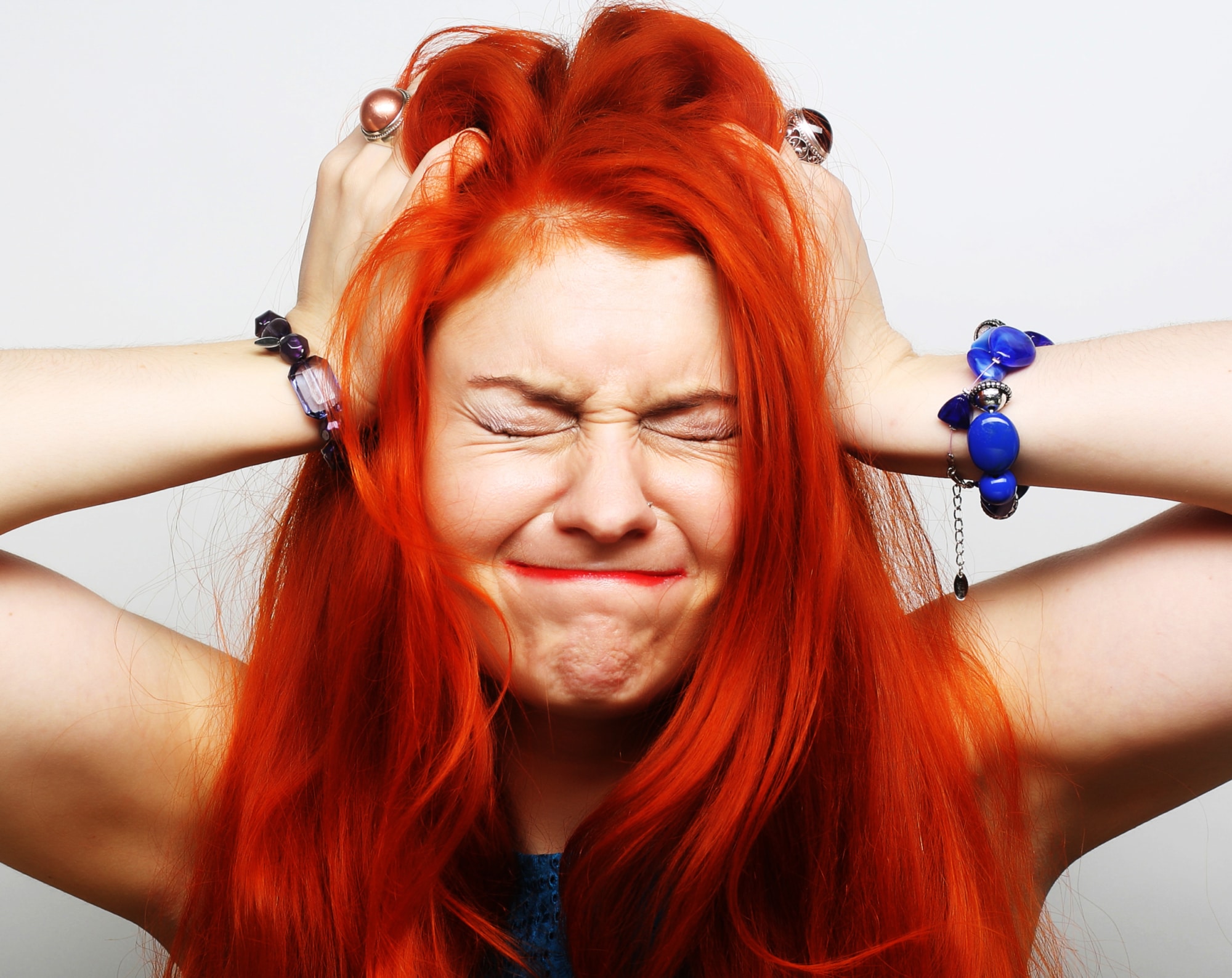 Person with long red hair and closed eyes presses hands to head, appearing frustrated or stressed, against a plain background.
