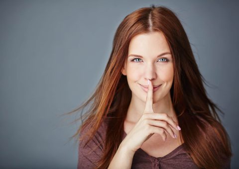 Woman with long brown hair holds her index finger to her lips, signaling "shh" or silence, against a plain grey background.