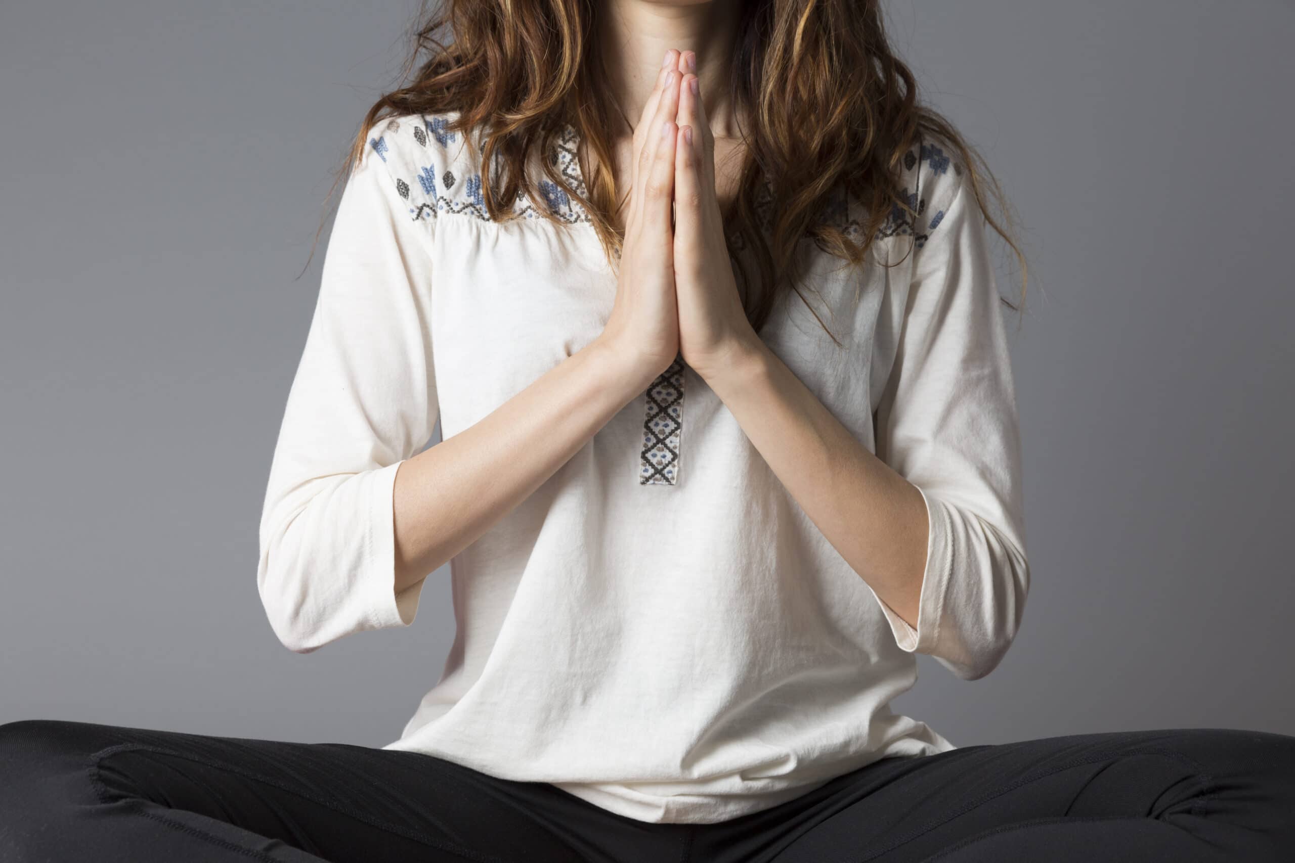 A person sits cross-legged with hands in a prayer position at chest level, wearing a white shirt with a patterned neckline.