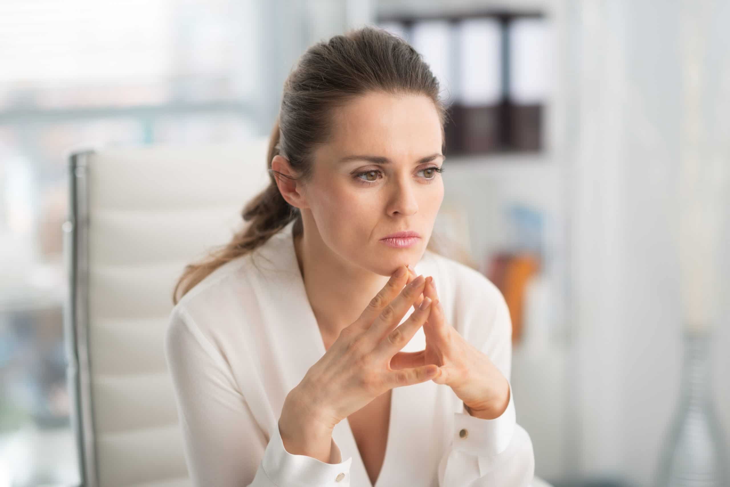 A woman sits in an office chair, looking thoughtful with her hands clasped near her face, wearing a white blouse.
