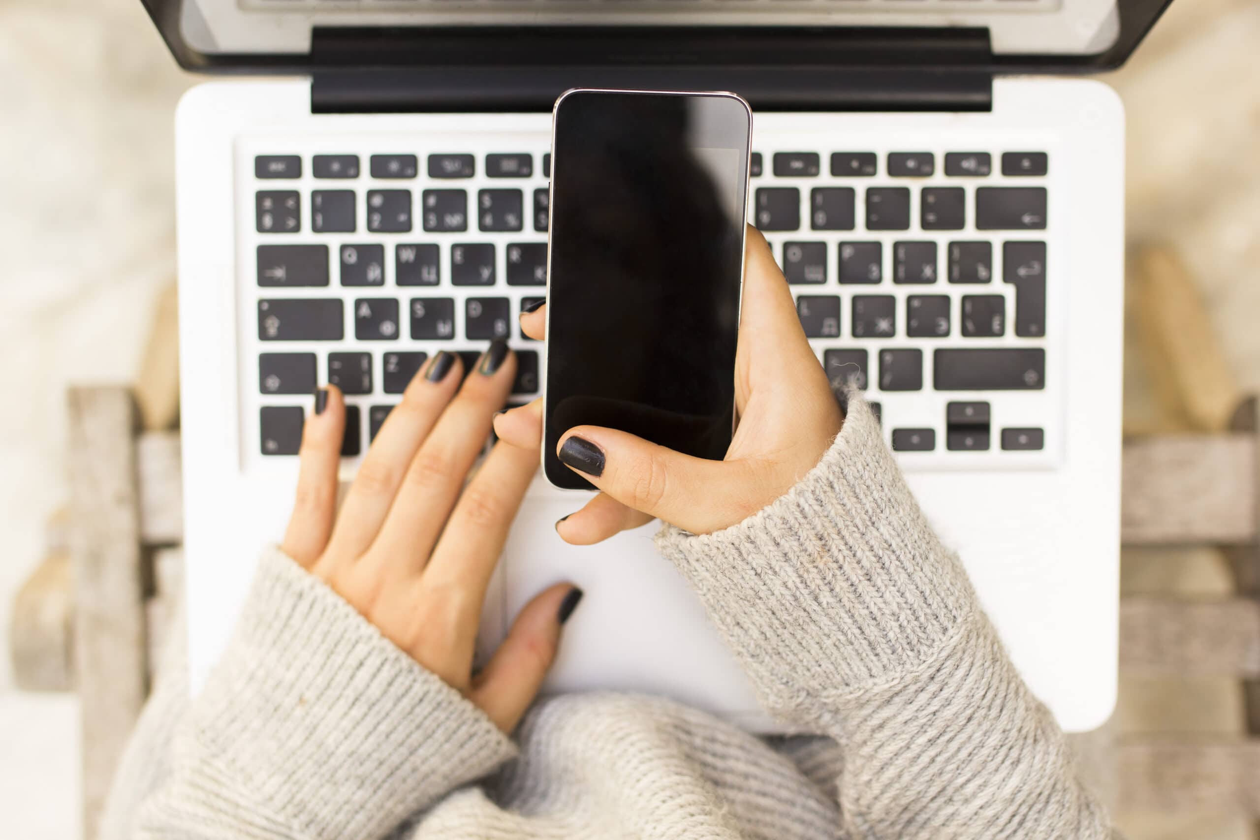 Person wearing a gray sweater using a laptop while holding a smartphone in one hand, photographed from above.