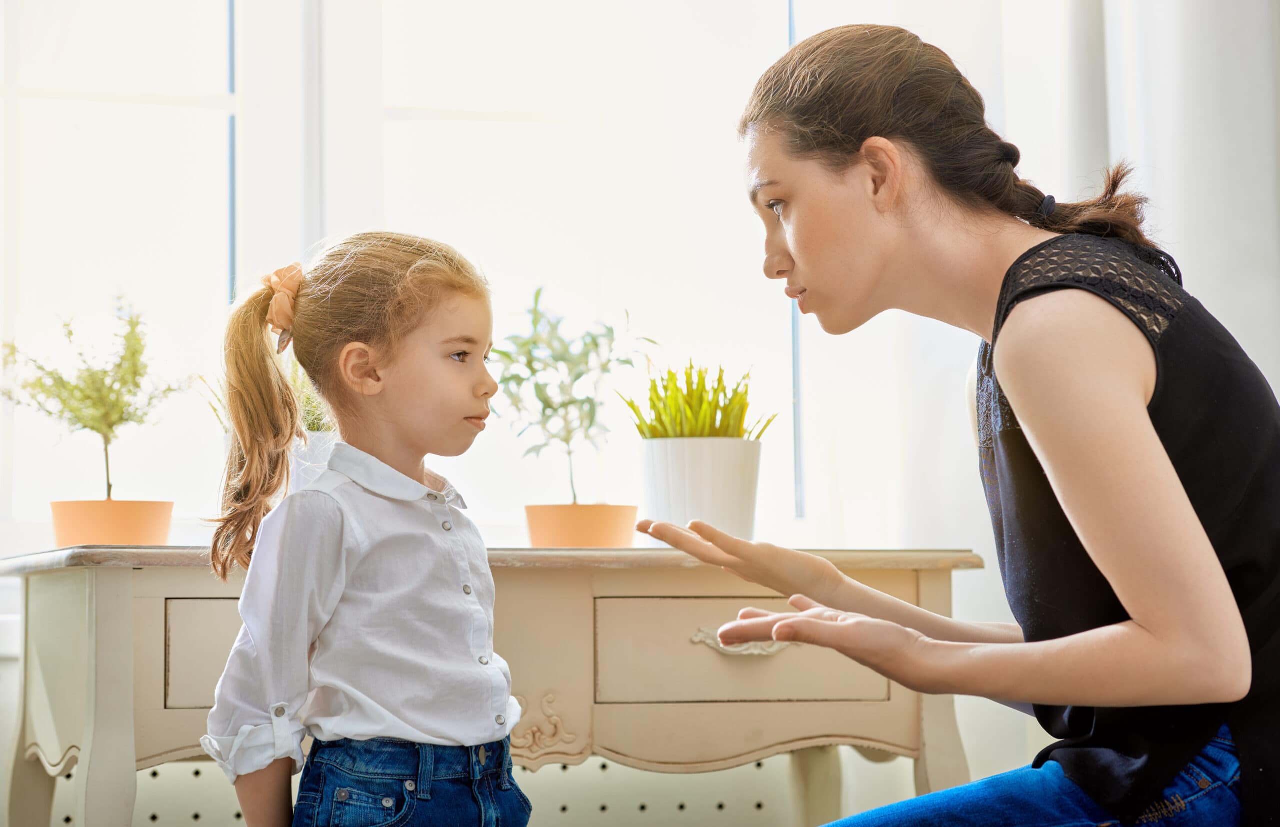 An adult woman is speaking seriously to a young girl indoors near a table with potted plants; the girl listens attentively.
