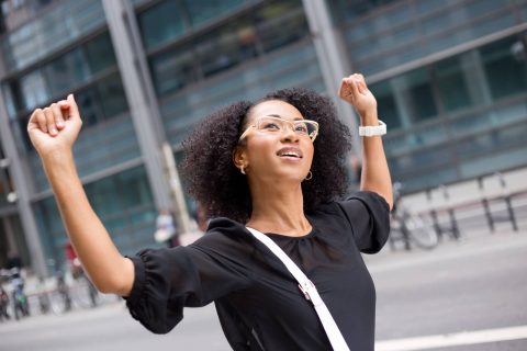A woman wearing glasses and a black top stands outdoors in a city with her arms raised, smiling and looking upward.