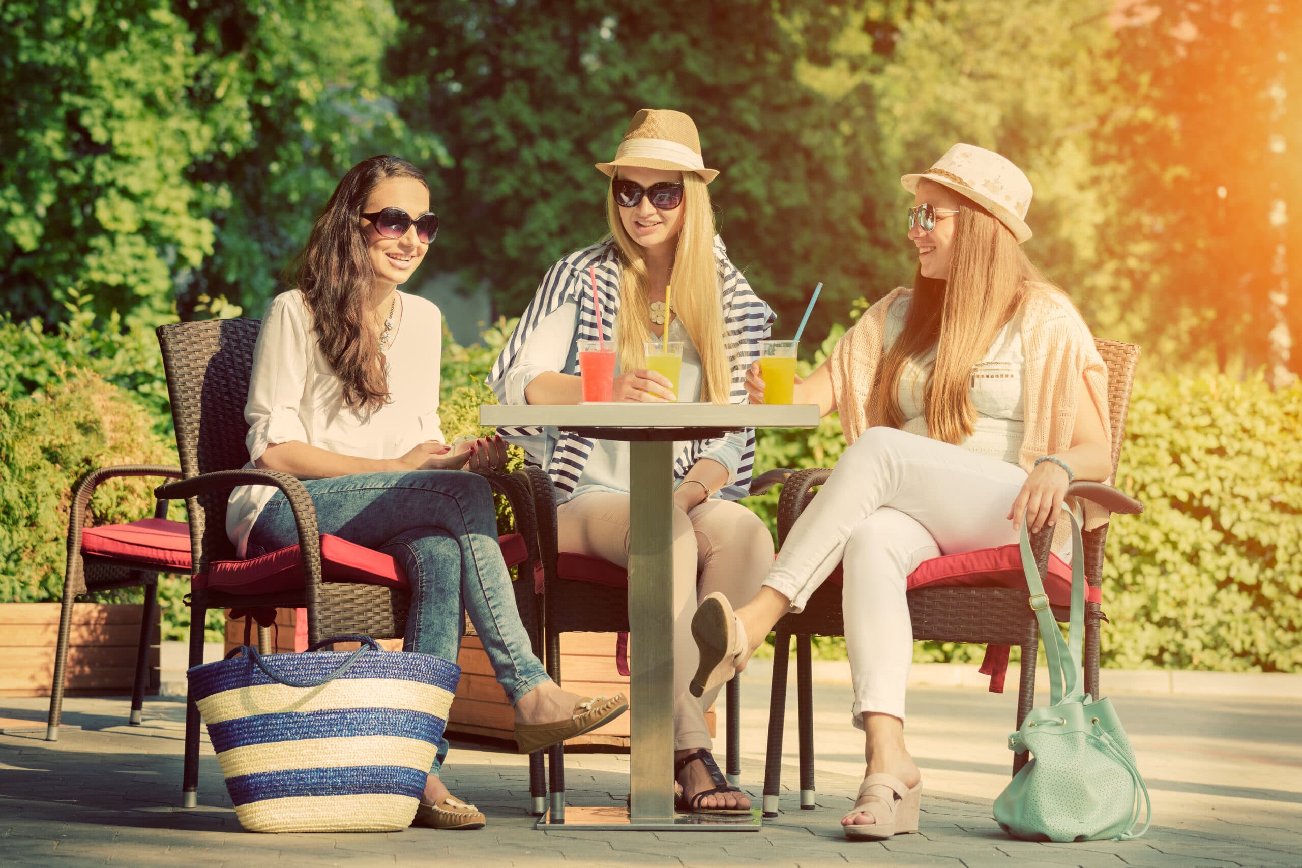 Three women sit at an outdoor cafe table, smiling and chatting, with drinks on the table and handbags on the ground, in a sunny setting with greenery in the background.