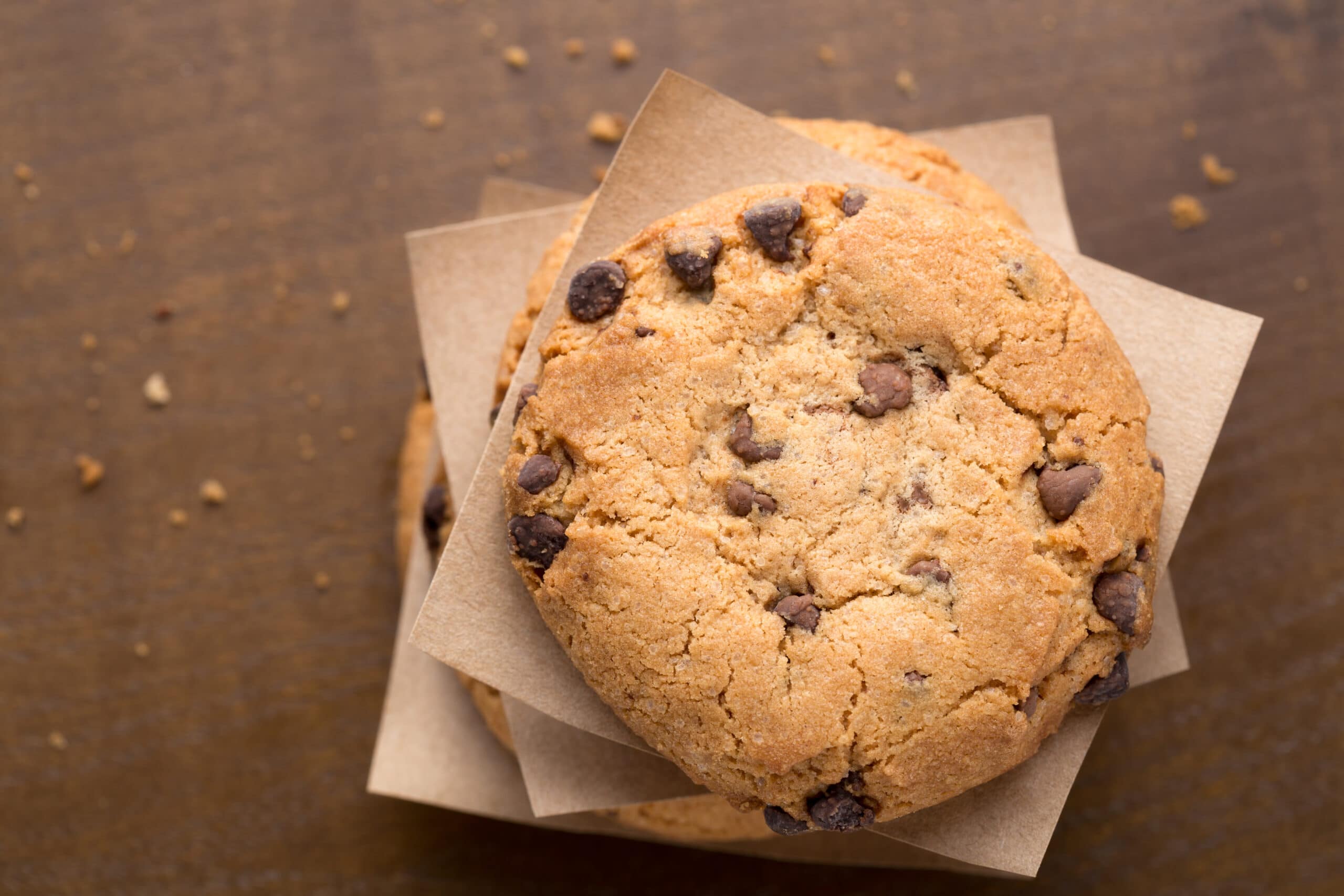 A stack of chocolate chip cookies separated by parchment paper, viewed from above on a wooden surface with a few crumbs scattered around.