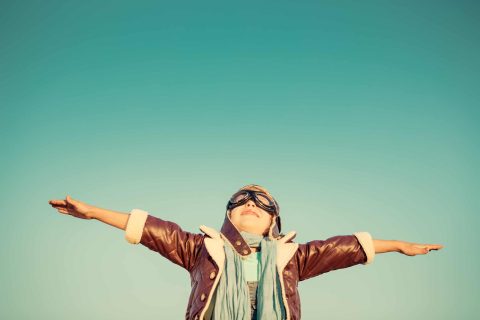 Child wearing aviator goggles and a leather jacket stands outdoors with arms outstretched under a clear sky.