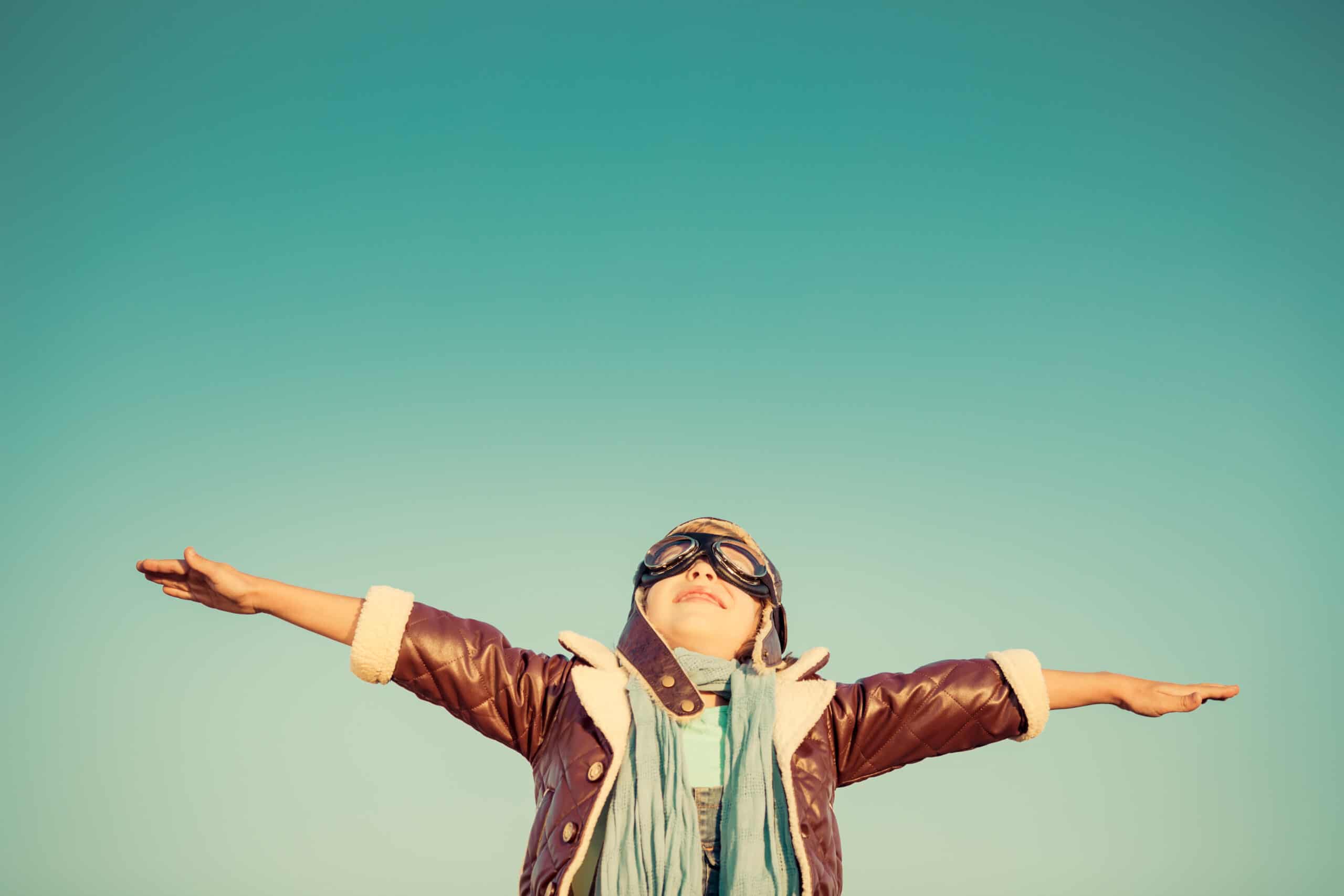 Child wearing aviator goggles and a brown jacket stands outside with arms outstretched against a clear blue sky.
