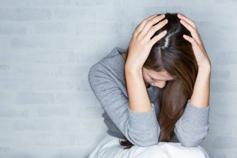 A woman sits with her head down and hands covering her head, appearing distressed, against a light-colored brick wall.