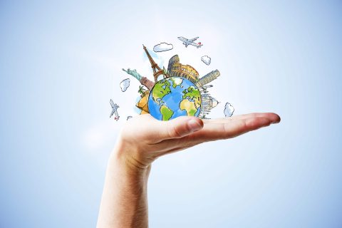 A hand holds a small illustrated globe with famous landmarks, airplanes, and clouds, symbolizing global travel and tourism.