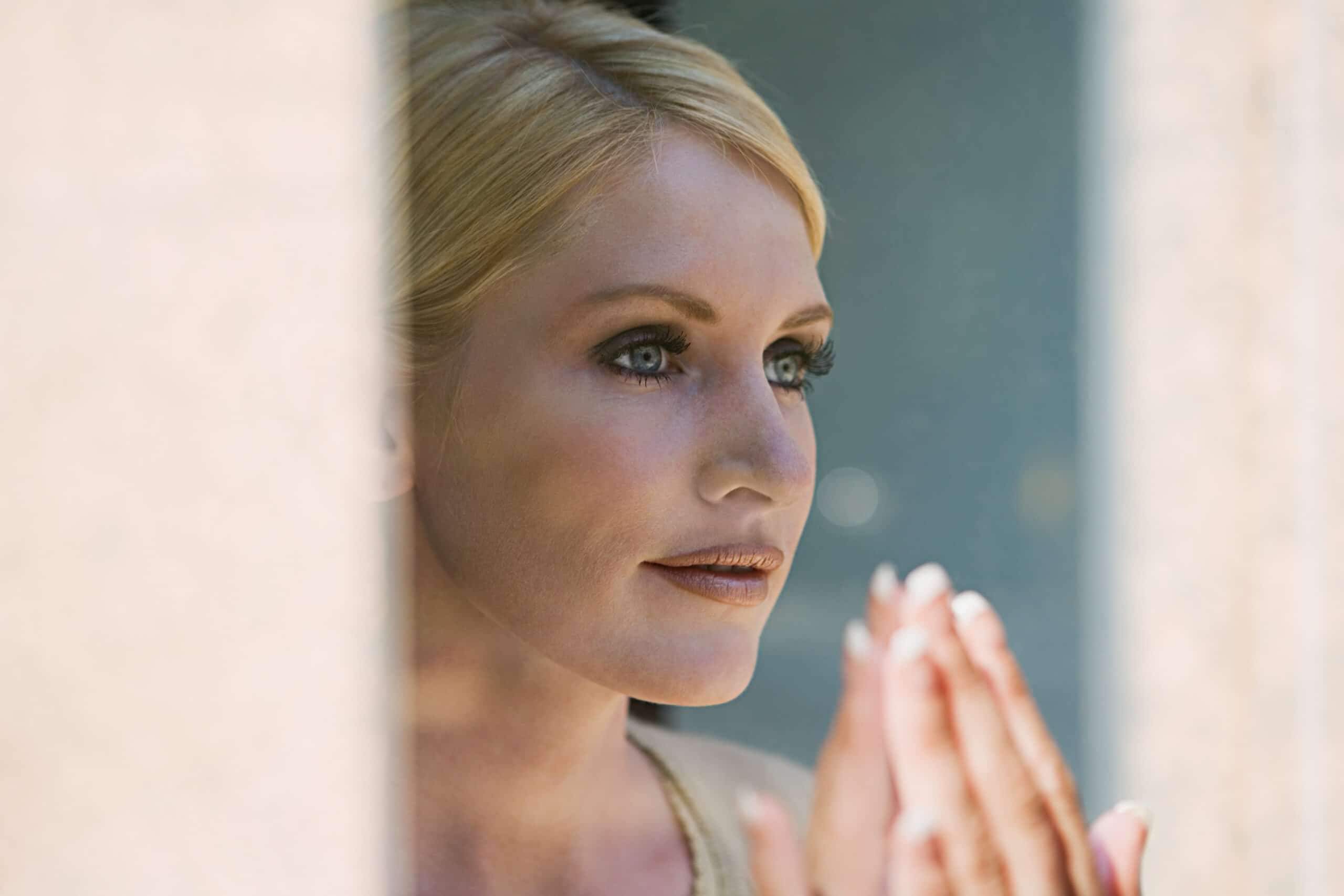 A woman with blonde hair and light eyes looks thoughtfully out of a window with her hands pressed together near her face.
