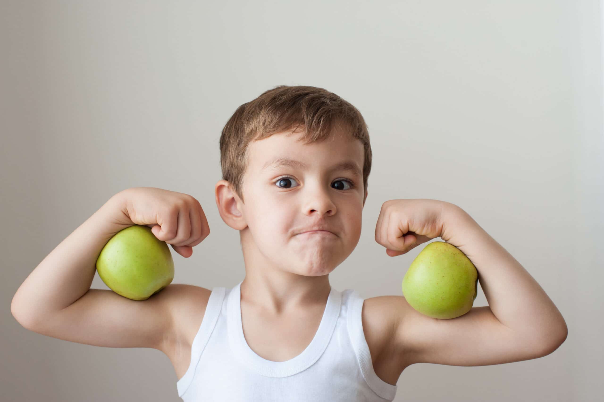 A young boy in a white tank top holds green apples at his biceps, flexing his arms and making a serious face against a plain background.