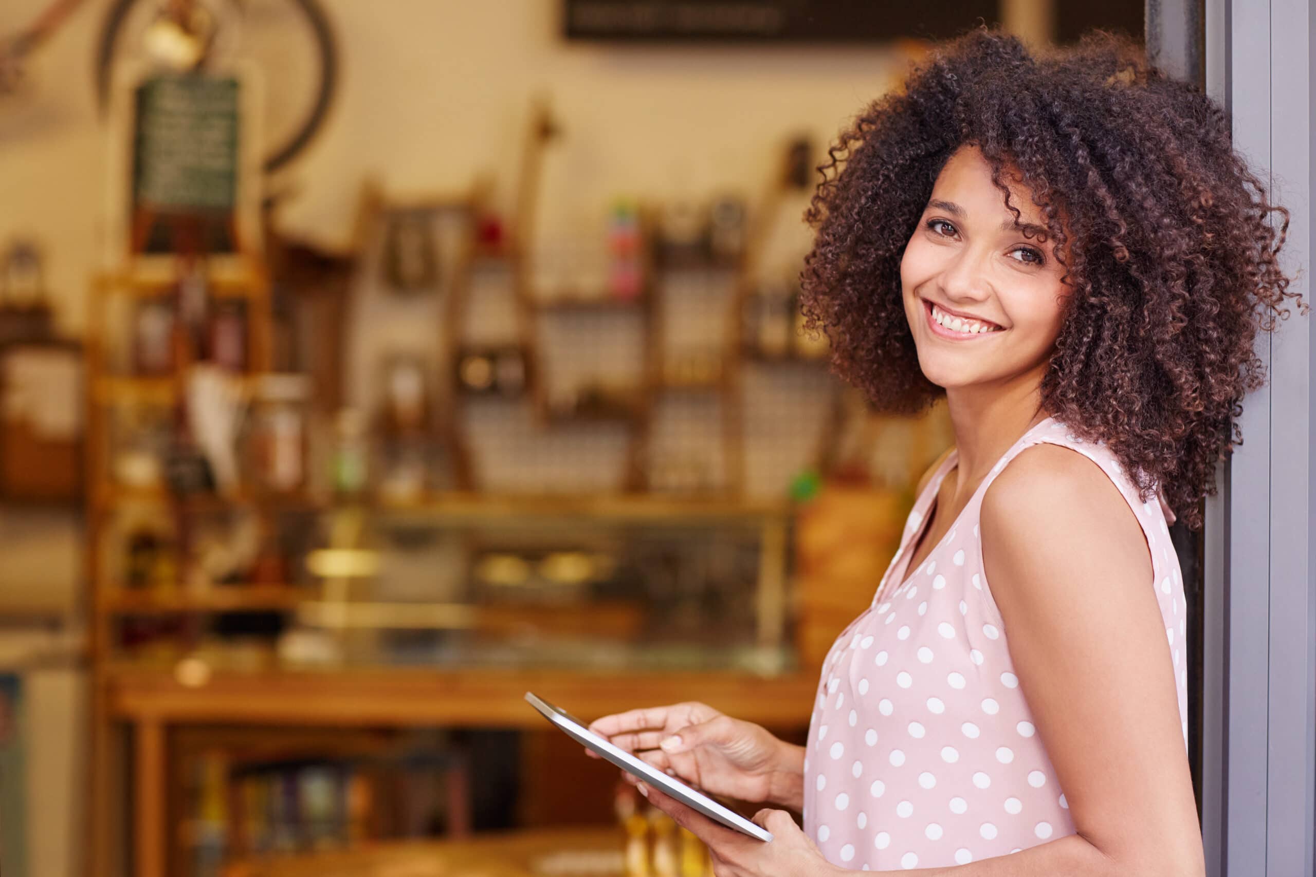A woman with curly hair wearing a pink polka dot blouse stands by a shop entrance, smiling and holding a tablet.