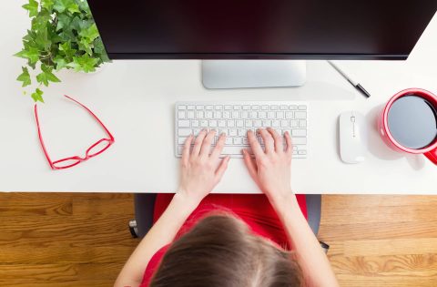 Person typing on a wireless keyboard at a white desk with a plant, red glasses, a cup of coffee, and a computer monitor.