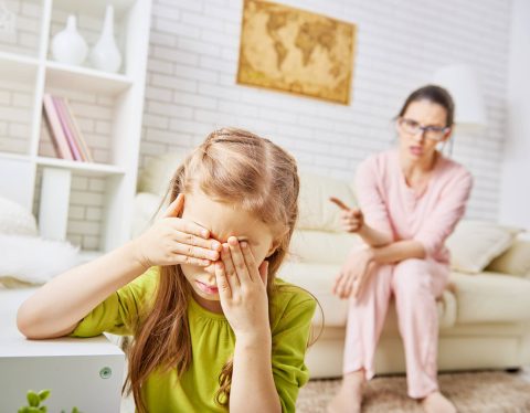 A young girl covers her face with her hands while a woman in the background points and appears to be scolding her in a living room.
