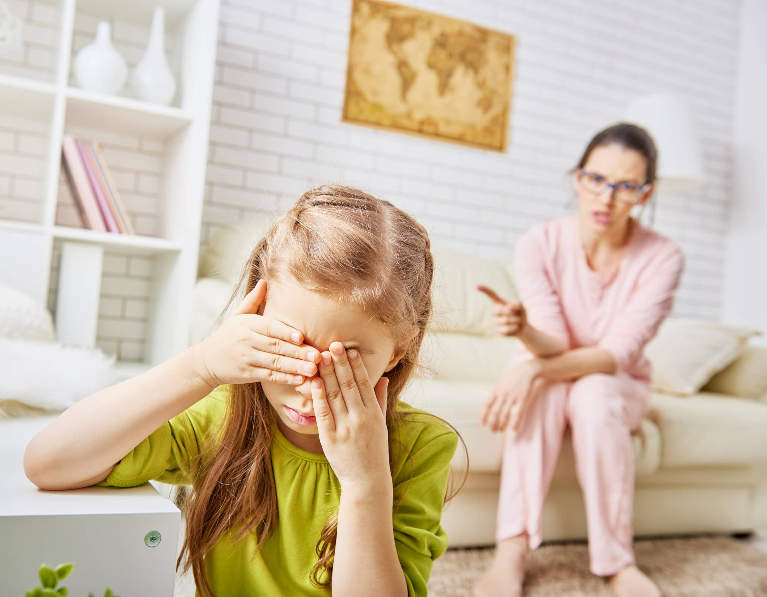 A young girl covers her face with her hands while a woman in the background points and appears to be scolding her in a living room.