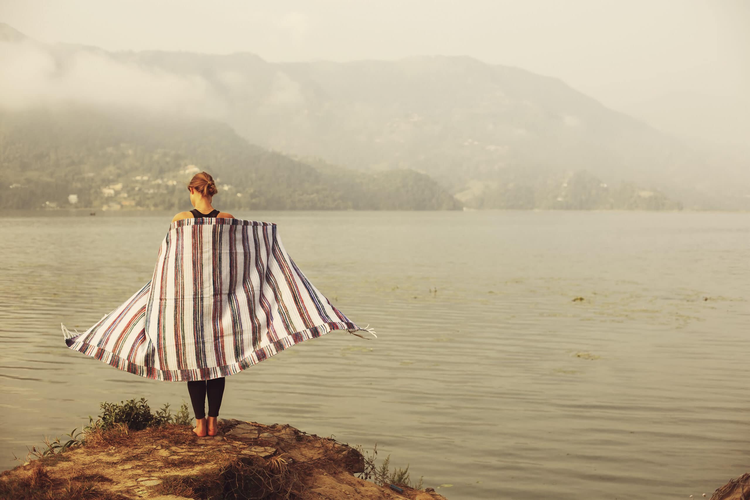 A person stands on a small hill by a lake, wrapped in a striped blanket, facing the water and distant mountains on a hazy day.