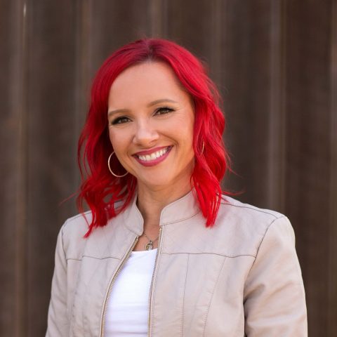A person with bright red hair, wearing a light-colored jacket over a white top, smiles in front of a dark, blurred background.