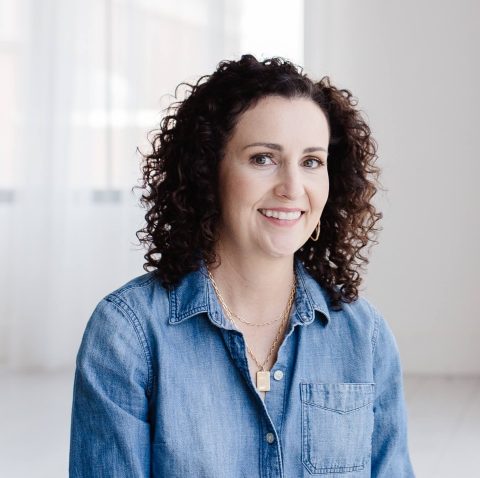 A woman with curly brown hair, wearing a denim shirt and gold jewelry, smiles at the camera in a bright, minimalistic room.